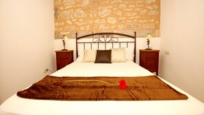 A neatly made bed with a brown blanket and a single red flower, two bedside lamps, and a stone-patterned wall above.