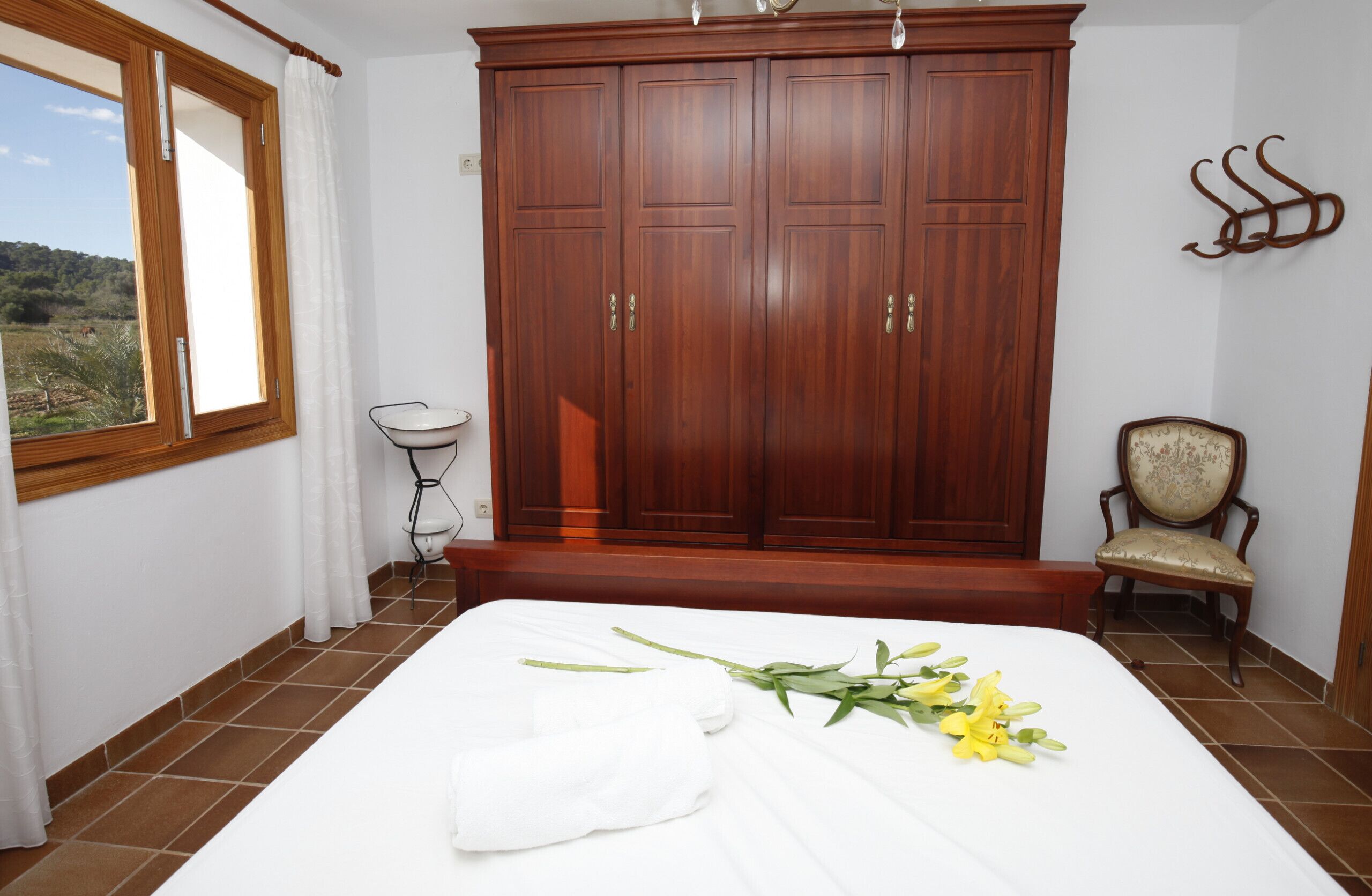A bedroom with a large wooden wardrobe, a bed with white linens, a chair, and a window with curtains.
