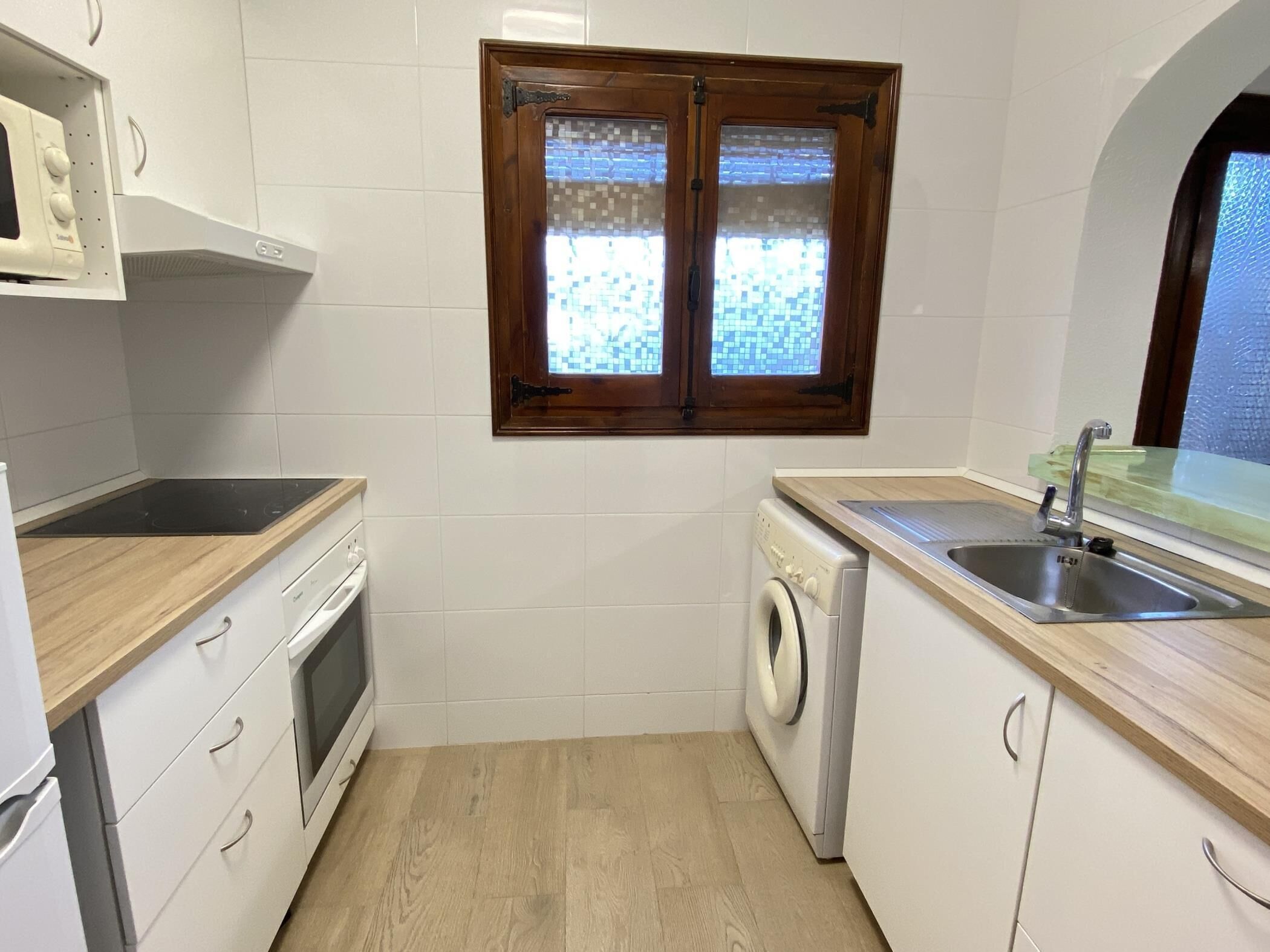 A compact kitchen with white cabinets, a built-in oven, a washing machine, and a window with a wooden frame.