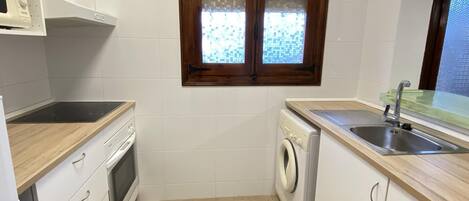 A compact kitchen with white cabinets, a built-in oven, a washing machine, and a window with a wooden frame.