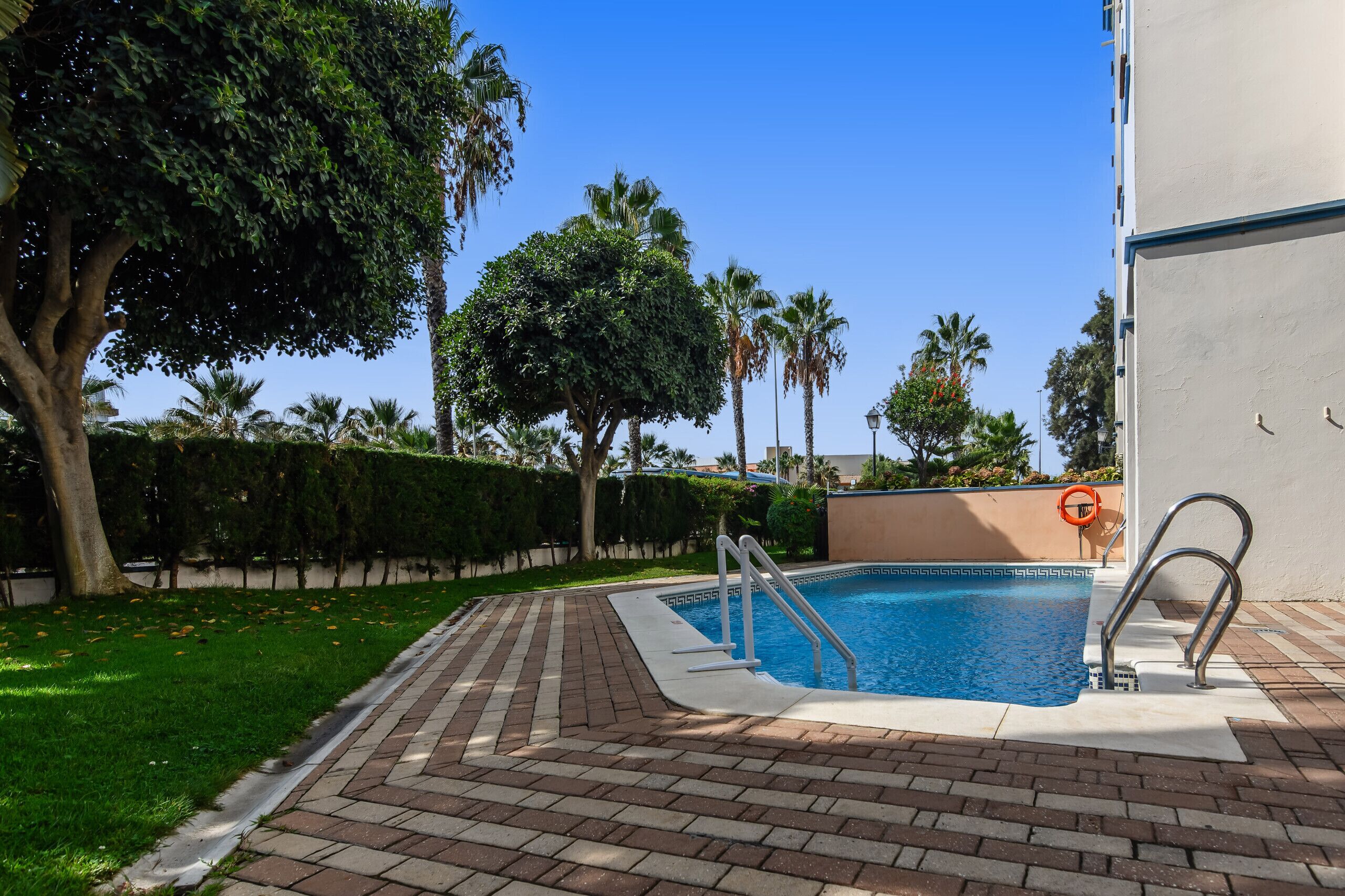 Una piscina con una escalera metálica, rodeada por un patio de ladrillo y vegetación.