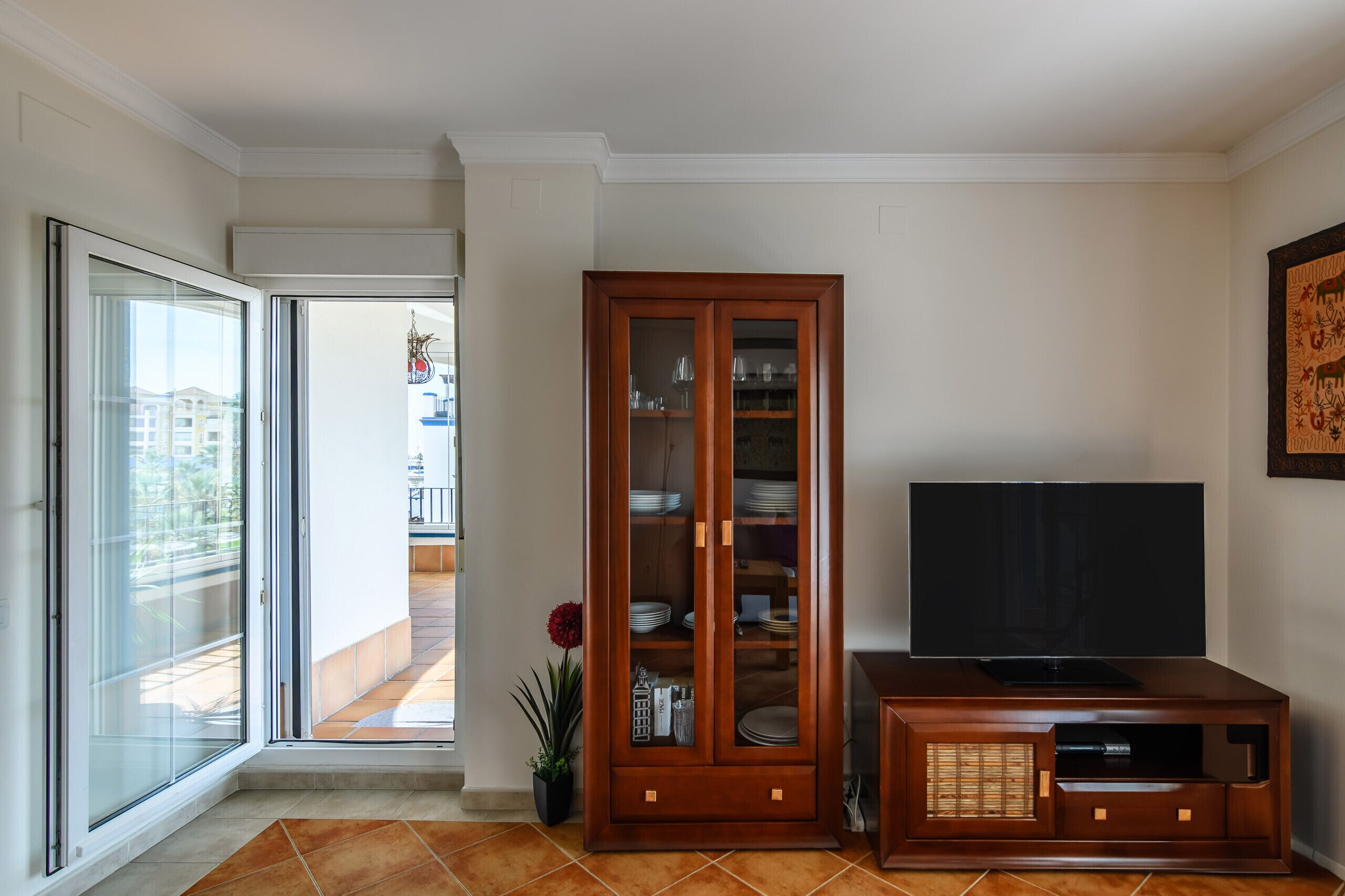 Un salon doté d’une porte-fenêtre coulissante donnant sur un balcon, d’un meuble en bois avec des portes en verre et d’un téléviseur à écran plat posé sur un meuble en bois.