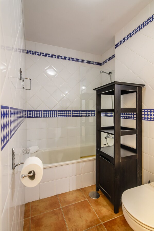 Un baño con baldosas blancas, una bañera, un inodoro y un estante de madera.