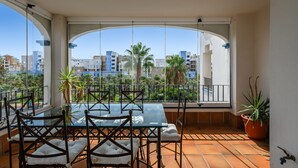 En balkon med et glasbord og stole, krukker med planter, og udsigt til bygninger og palmer.