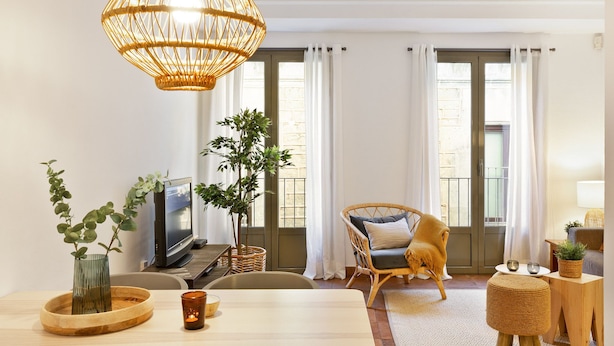 A cozy living room with a large woven light fixture, a television, wicker furniture, and a view of the city through large windows. - Columbus Apartment, - One Bedroom Apartment, Sleeps 2 (Barcelona)