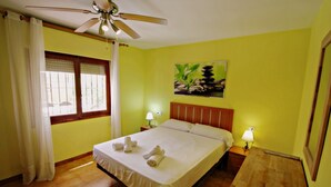 Une chambre d’hôtel avec un lit, deux lampes de chevet, un ventilateur de plafond, une fenêtre avec des rideaux et un tableau au mur.