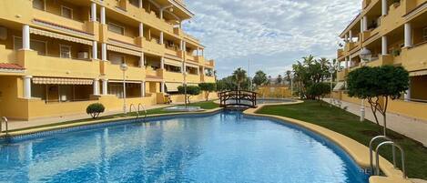 A swimming pool surrounded by yellow buildings with balconies, a bridge, and palm trees.