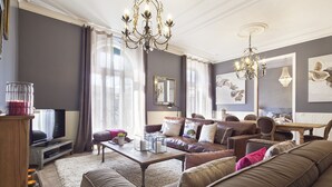 Ein modernes Wohnzimmer mit einer großen braunen Ledersofa, einem Holztisch und einem Fernseher auf einem Schrank. Zwei Kronleuchter, ein großes Fenster mit Vorhängen und ein Gemälde an der Wand sind ebenfalls zu sehen.