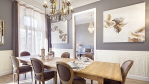 Ein Esszimmer mit Holztisch, sechs Stühlen und einer Chandelierleuchtre. An den Wänden hängen zwei große Gemälde und ein Fenster mit Vorhängen.