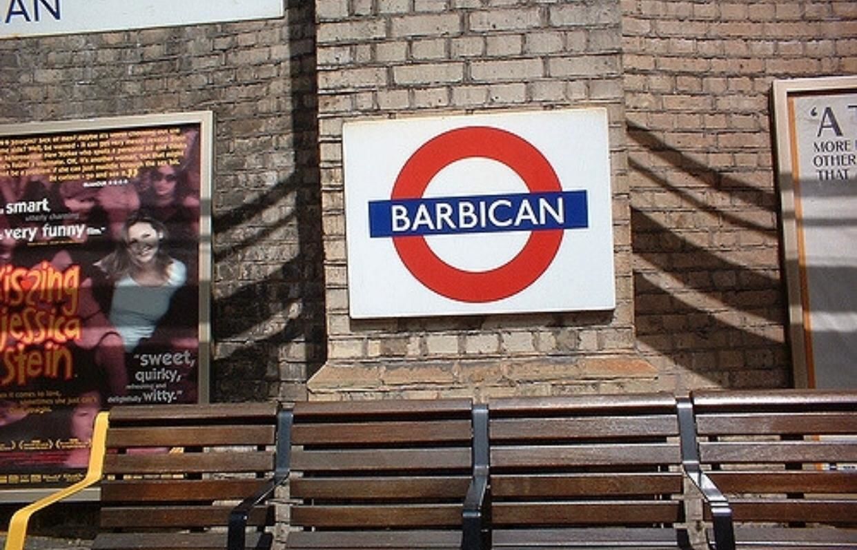 A sign for 'BARBICAN' with a red circle and blue bar design, mounted on a brick wall.