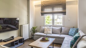 Un salon moderne avec un téléviseur à écran plat, une table basse en bois sur laquelle sont disposés des fruits, un canapé gris et une fenêtre avec des stores.
