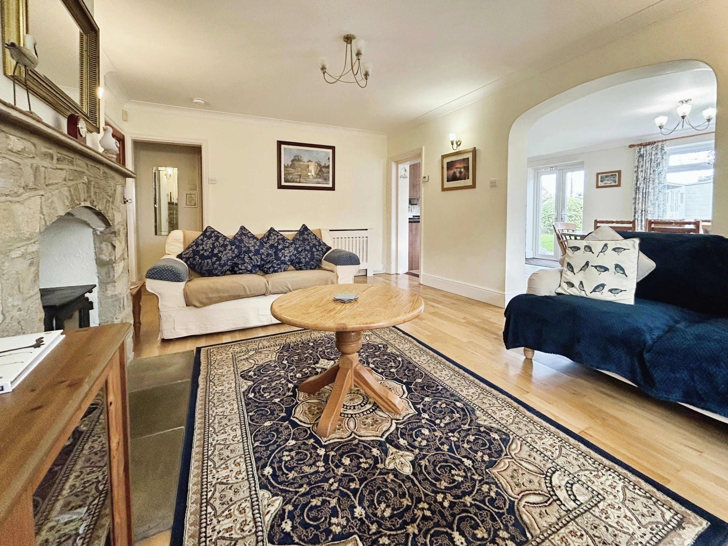 A living room with a stone fireplace, a round wooden coffee table, a patterned rug, a beige sofa with blue pillows, and a dark blue sofa with bird-patterned pillows.
