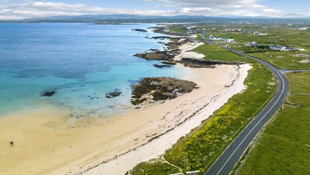 Luftaufnahme einer Küstenregion mit Sandstrand, klarem, blauem Wasser und einer sich an der Küste entlang schlängelnden Straße.