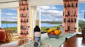 Ein Essbereich mit Tisch für zwei, einer Weinflasche und einer Obstschale. Der Raum verfügt über große Fenster mit Vorhängen und bietet Blick auf eine Landschaft mit Wasser und Hügeln.
