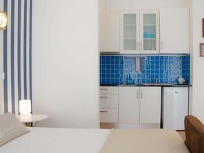 Une pièce de taille modeste comprenant un lit, une petite table, un coin cuisine avec des meubles blancs et un plan de travail bleu recouvert de carreaux.