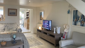 Ein gemütliches Wohnzimmer mit Fernseher, einer Couch, einem gemusterten Sessel und einem Essbereich, der durch eine offene Tür sichtbar ist.