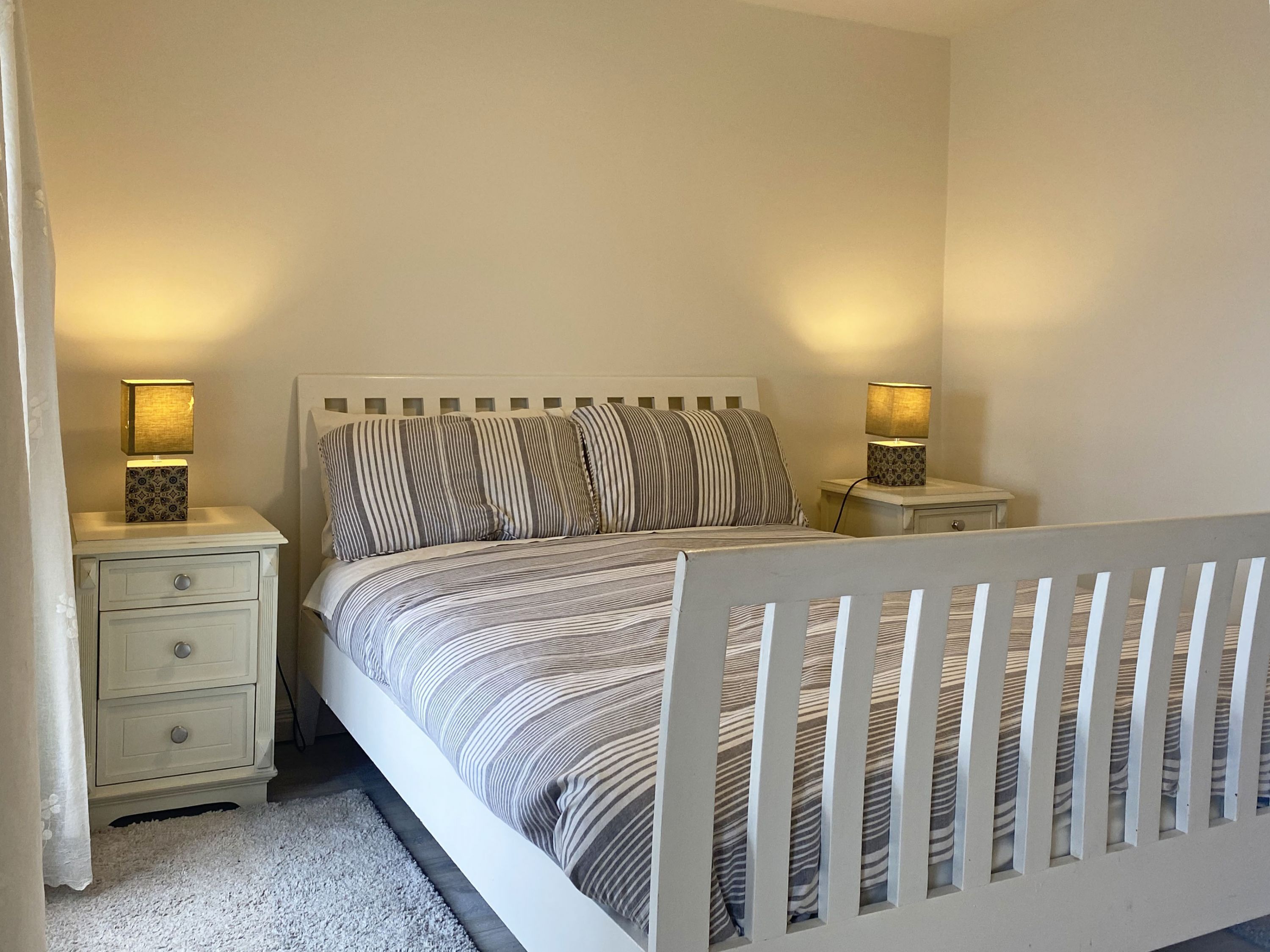 A bedroom with a bed, two bedside tables, a nightstand, a crib, and a white curtain.