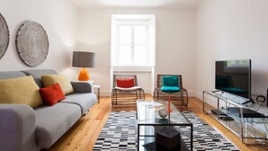 Una sala de estar moderna con un sofá gris, sillas de mimbre, una mesa de centro de vidrio y un televisor sobre un mueble.