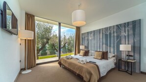Een hotelkamer met een groot bed, twee nachtkastjes, een groot raam met gordijnen, en uitzicht op bomen en een hek.