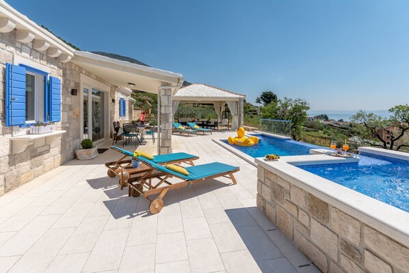 Zona de piscina con sillones de descanso, un área de estar techada y vistas al mar.