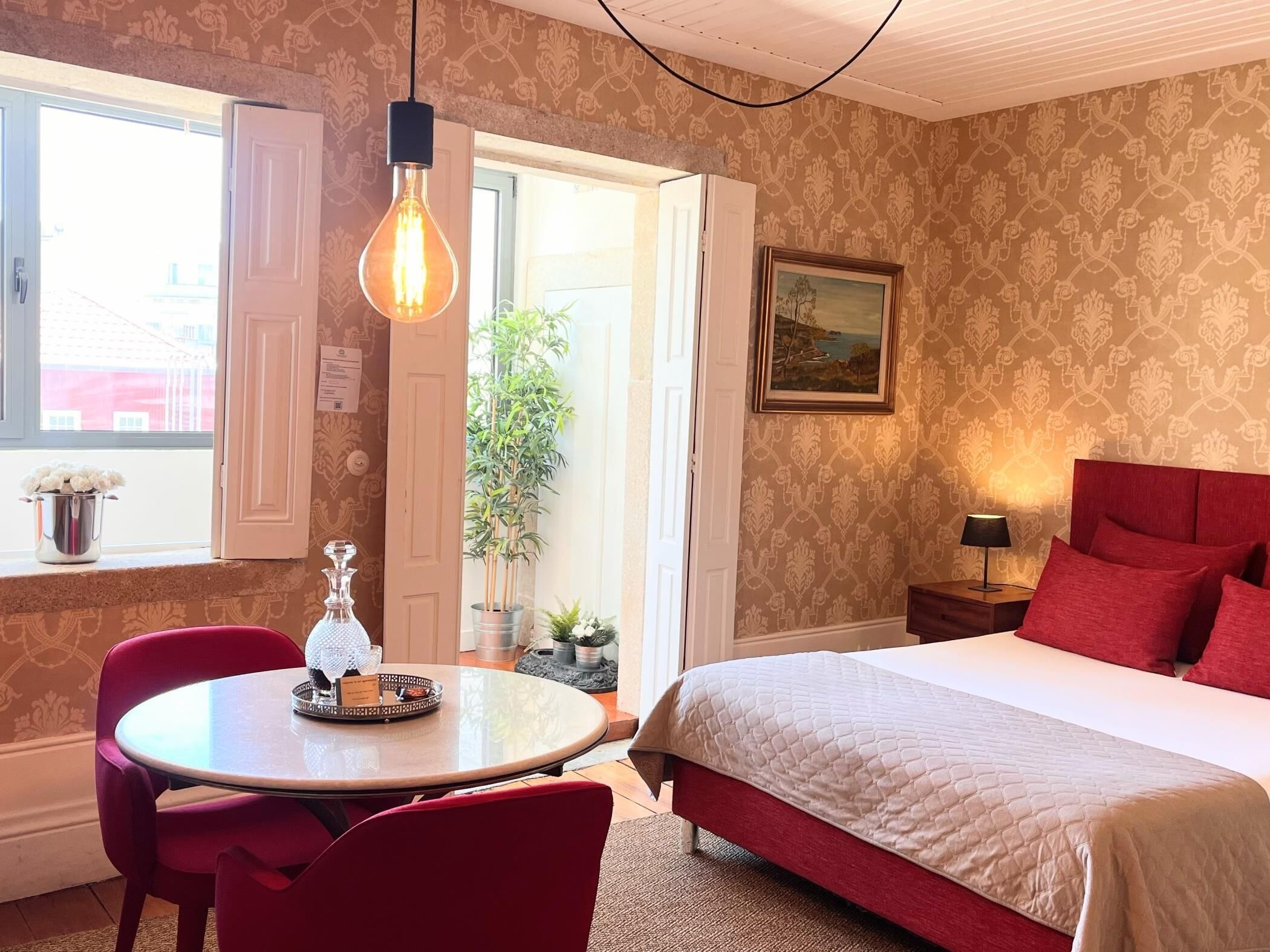 A hotel room with a bed, a round table with chairs, a potted plant, a painting on the wall, and a window with a view.