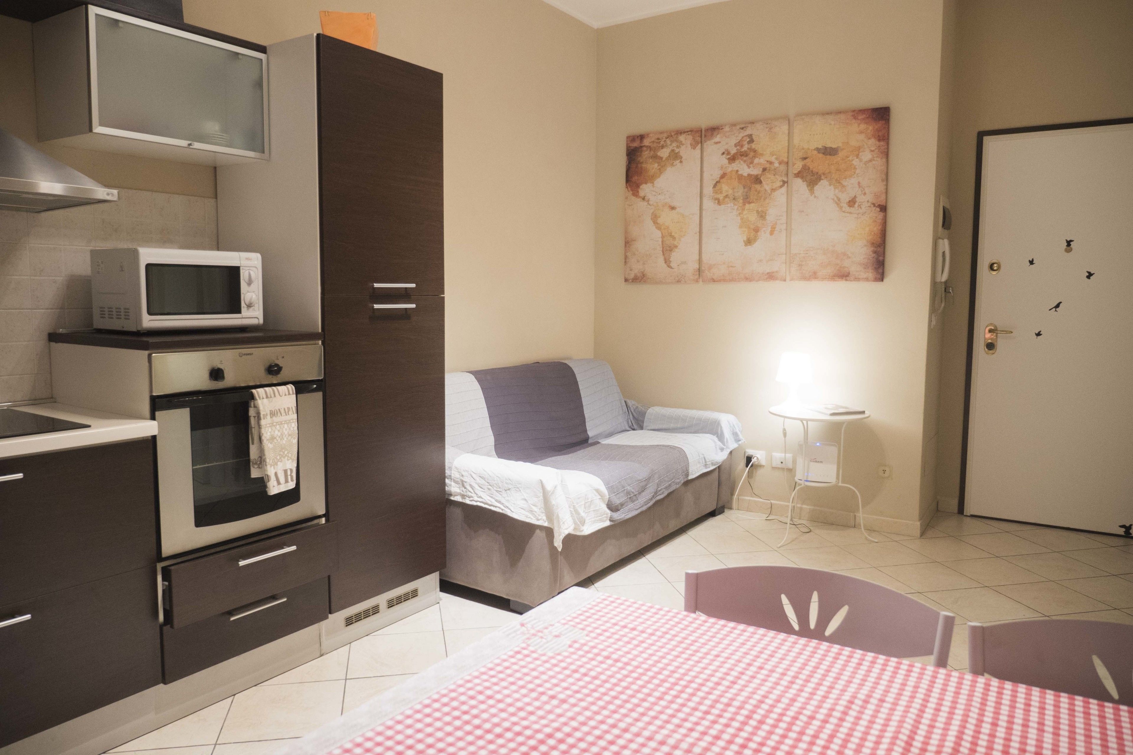 A compact hotel room with a kitchenette, a bed with a gray blanket, a small bedside table with a lamp, and a world map artwork on the wall.