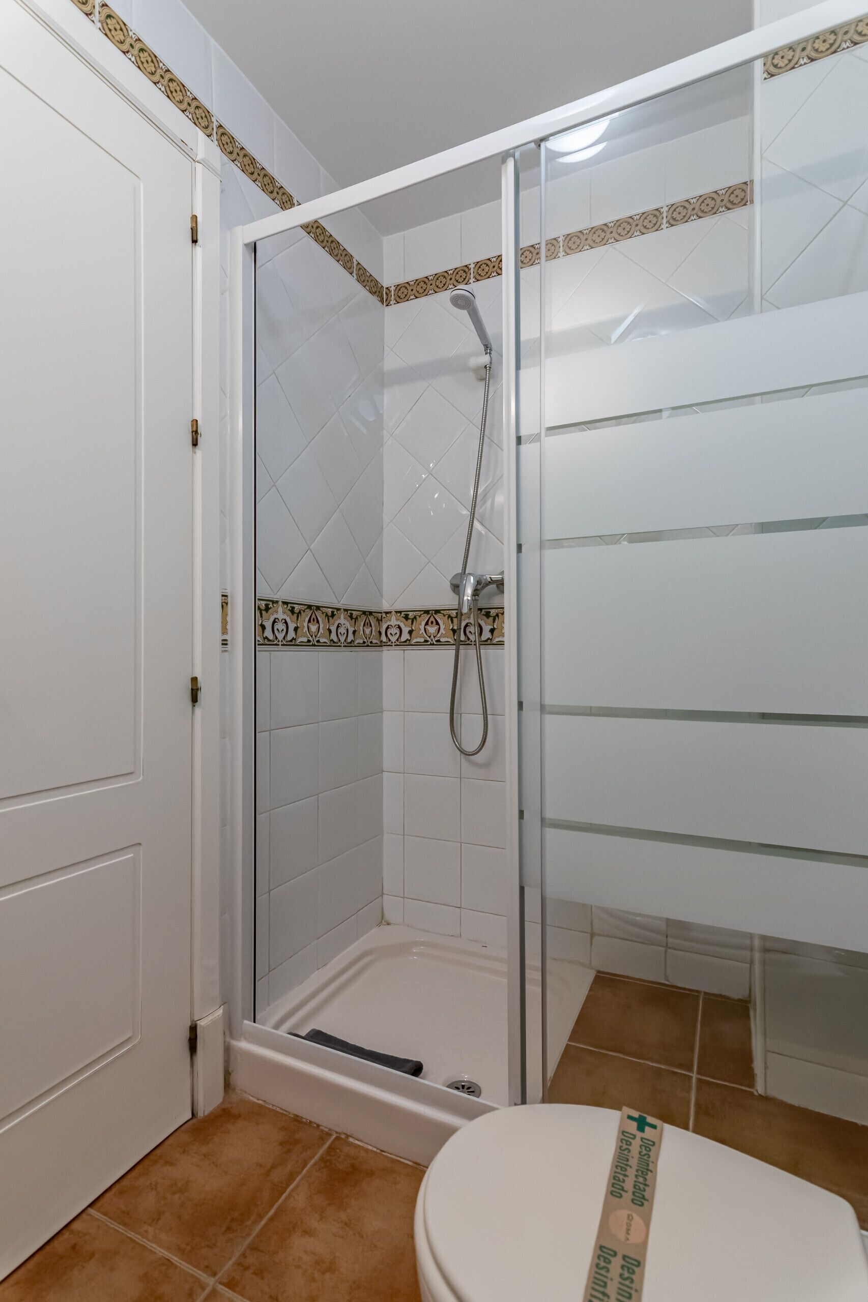 A bathroom with a glass-enclosed shower, white tiles, and a toilet with a green sign.