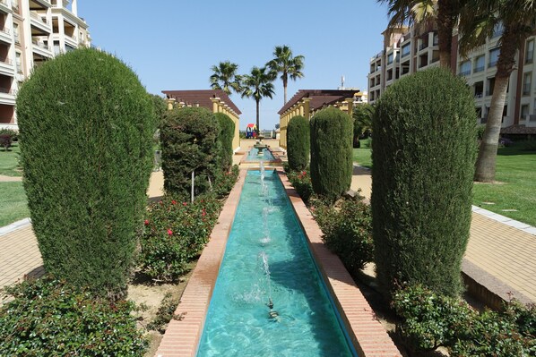 A long, narrow pool with water jets, flanked by neatly trimmed hedges and a brick pathway.