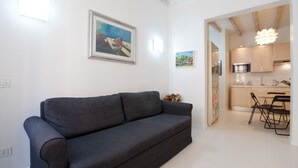 A modern living room with a grey sofa, a dining area with a table and chairs, and a kitchen visible through an open doorway.