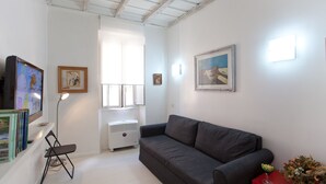 A living room with a grey sofa, a small table with a vase, a TV, and a window with blinds.