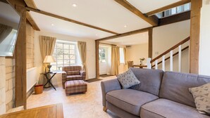 A cozy living room with a sofa, a plaid armchair, and a lamp. A staircase with wooden beams leads to another room. - A sudeley castle cottage that sleeps 4 guests  in 2 bedrooms (Cheltenham)