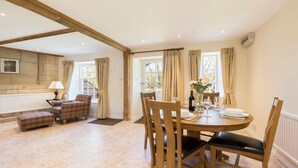 A dining area with a round wooden table set for four, flanked by two wooden chairs with black cushions. A plaid armchair and a small ottoman are positioned near a window with patterned curtains. - A sudeley castle cottage that sleeps 4 guests  in 2 bedrooms (Cheltenham)