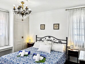 A hotel room with a bed, a chandelier, a wardrobe, and a nightstand with a lamp.