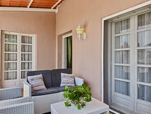 Una habitación con un sofá gris, muebles de mimbre blanco, una planta en maceta y un ventanal con cortinas.