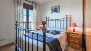 A bedroom with a bed, a nightstand, a lamp, a window with curtains, and a painting on the wall. - Playa Verde - Punta del Moral Beach PPHV31P26 (Punta del Moral)