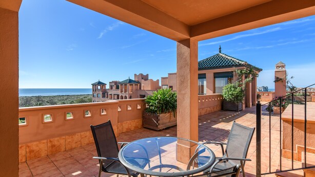 A balcony with a glass table and chairs, potted plants, and a view of the sea and buildings. - Playa Verde - Punta del Moral Beach PPHV31P26 (Punta del Moral)