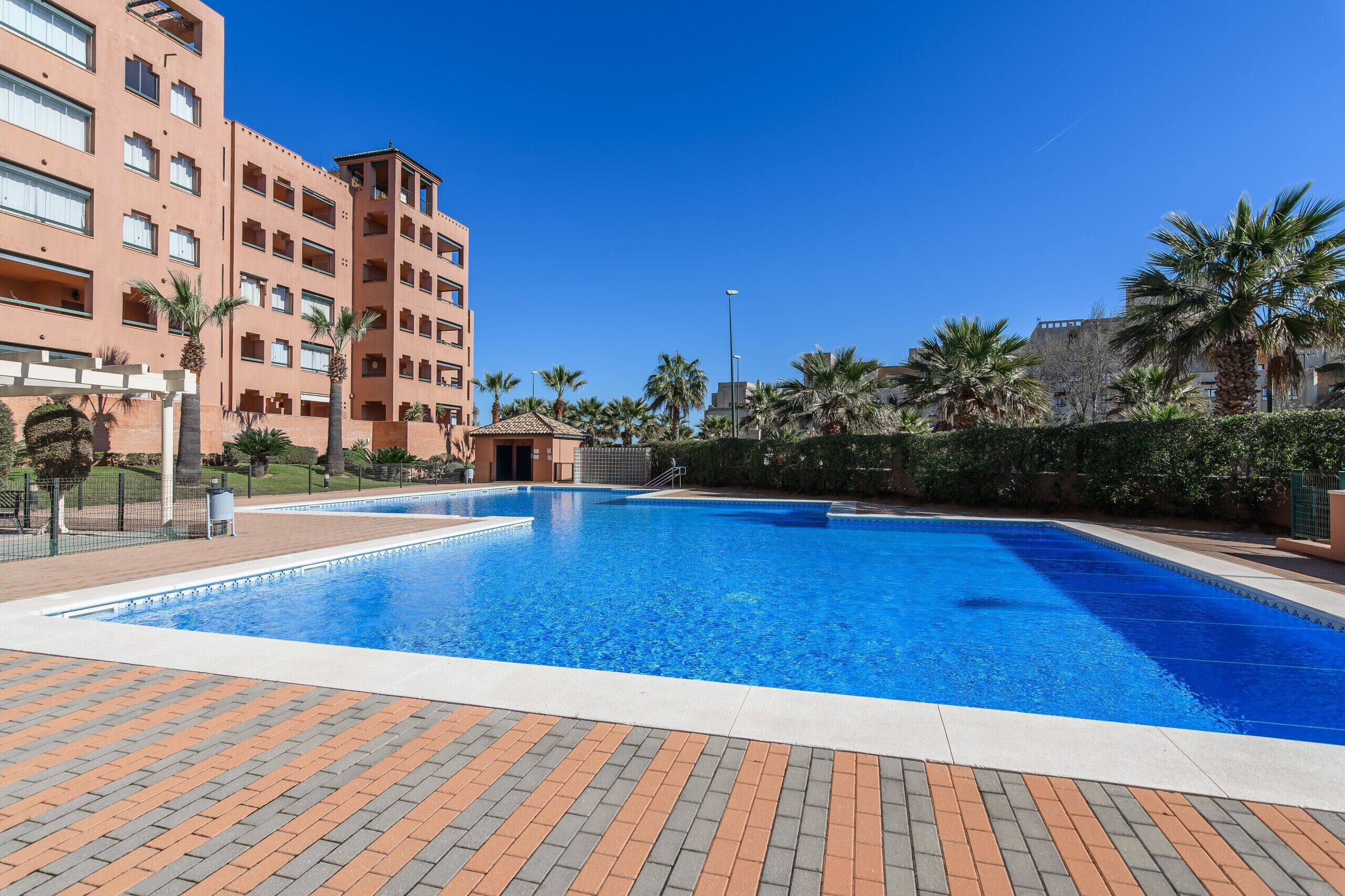 A swimming pool with a tiled deck, surrounded by palm trees and a multi-story building with balconies.