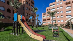 A playground with a slide and swings, surrounded by a well-maintained lawn and palm trees, with a multi-story building in the background. - Playa Verde - Punta del Moral Beach PPHV31P26 (Punta del Moral)