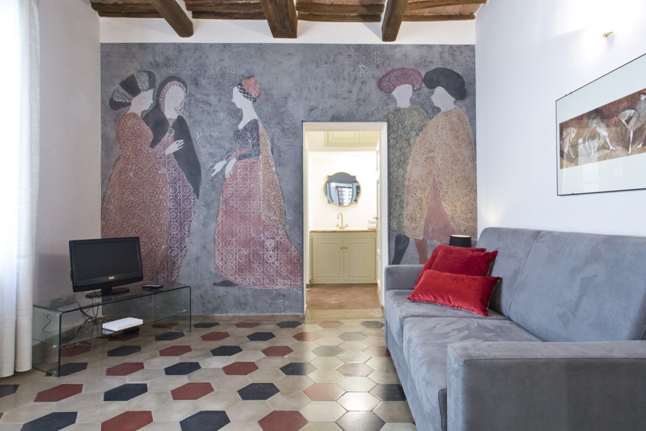 Een woonkamer met een groot muurschildering, een grijze bank met rode kussens, een glazen salontafel en een flatscreen televisie.