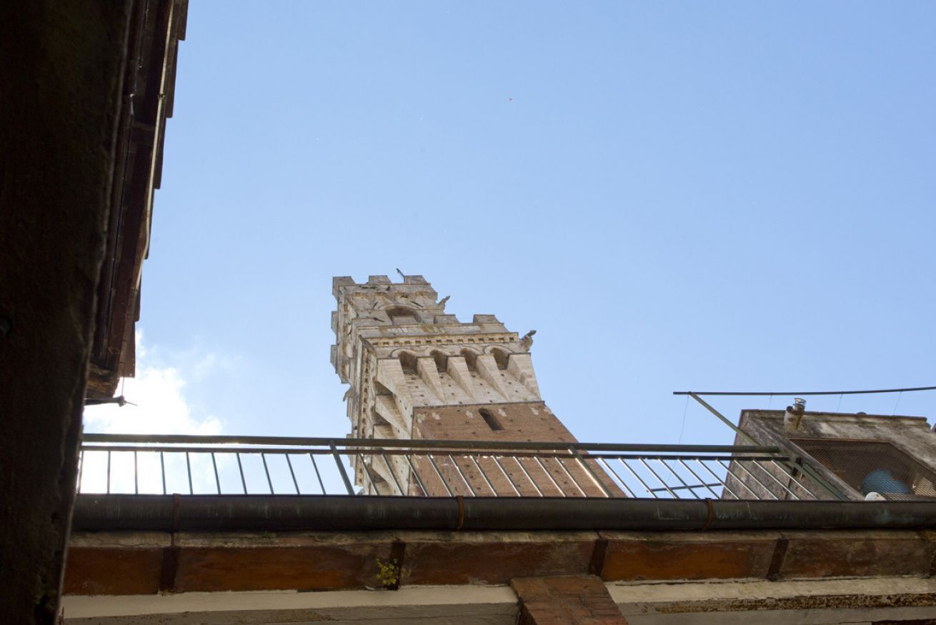Ein hoher, historischer Turm mit Zinnen, betrachtet von einem tiefer gelegenen Gebäude mit Metallgeländer.