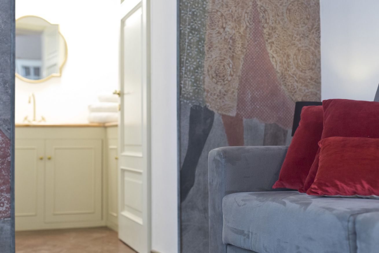 Una stanza con un divano grigio, cuscini rossi, una porta bianca e un muro con un parziale scorcio di un tessuto decorato.