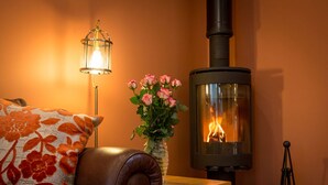 Una habitación acogedora con un sofá de cuero, un cojín floral, un jarrón con flores rosas, una lámpara de pared y un estufa de leña.