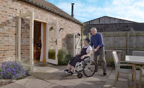 A wheelchair-accessible outdoor patio with a brick building, a wooden fence, and a table with chairs.