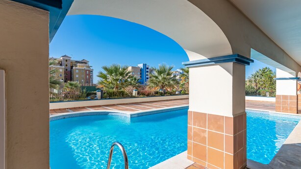 Une piscine à l’eau d’un bleu cristallin, entourée de murs recouverts de carreaux et de palmiers, avec des bâtiments en arrière-plan.