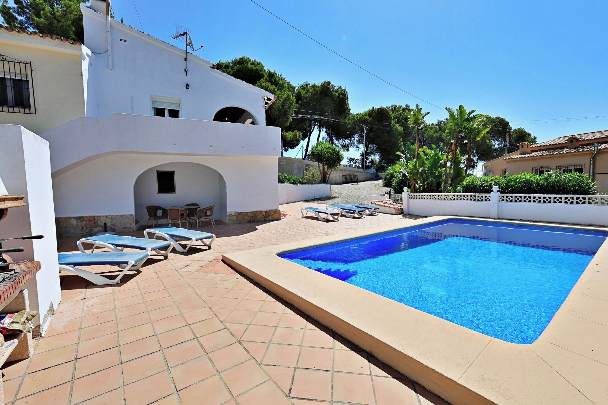 A swimming pool with lounge chairs, a covered seating area, and a white building with a tiled patio.