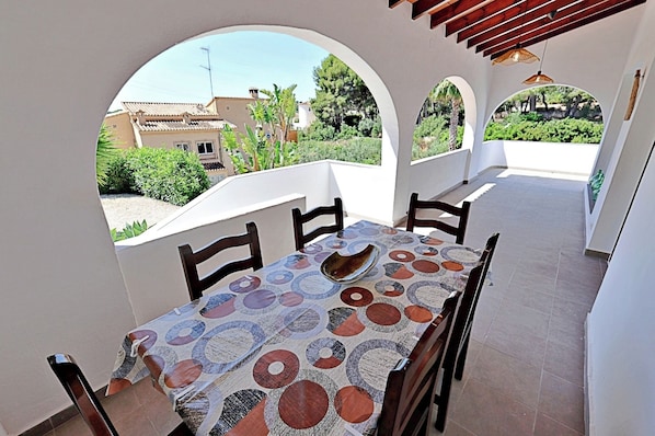 A covered outdoor dining area with a table and chairs, large arched windows, and a view of a residential area.