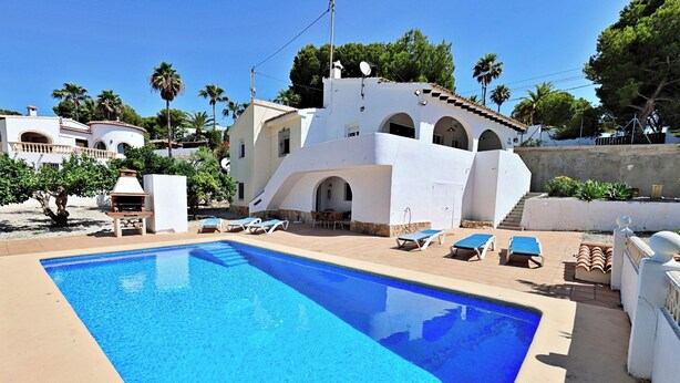 Une piscine avec des chaises longues, un barbecue et un bâtiment blanc orné d’arches.