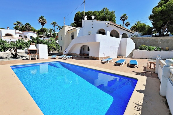 A swimming pool with lounge chairs, a barbecue, and a white building with arches.