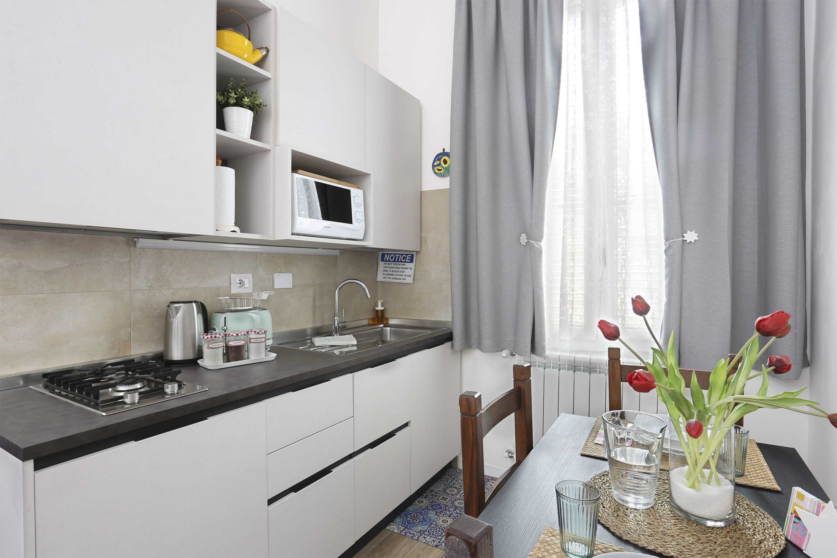 Cocina compacta con armarios blancos, un microondas, un hervidor de agua y un lavabo. Hay una mesa de comedor con un florero de tulipanes rojos y un vaso de agua.