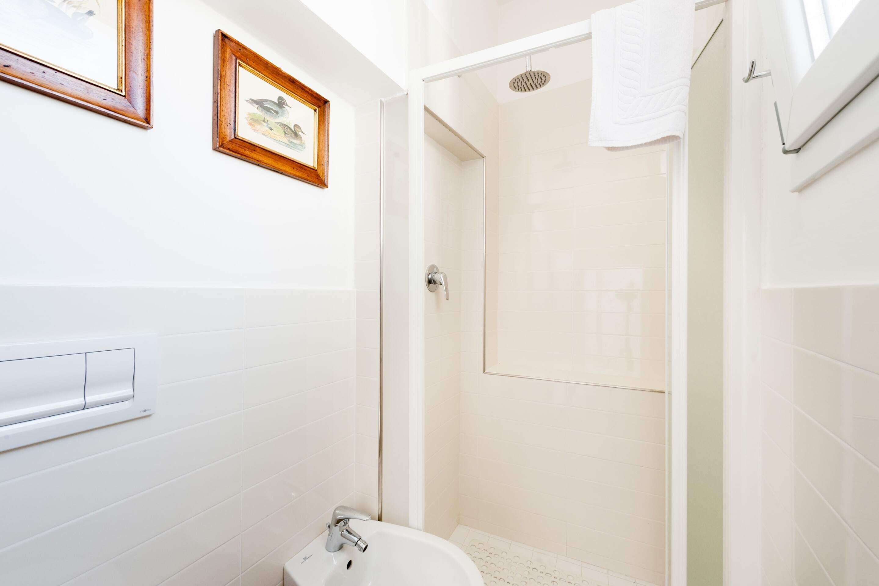 Een badkamer met witte tegels, een witte wastafel en een douche. Twee ingelijste foto's hangen aan de muur.